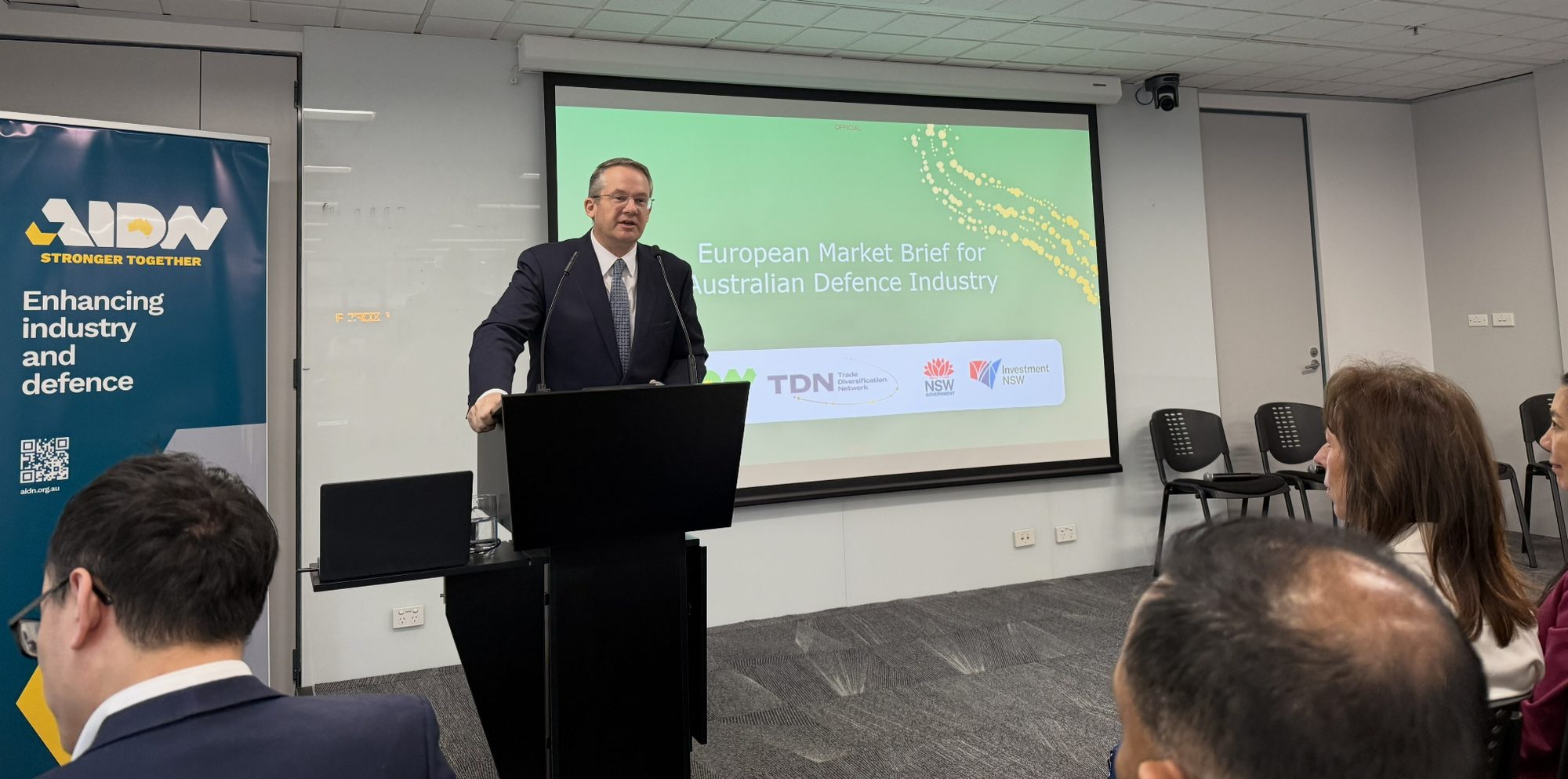 The host at the lectern opening the European Market Brief for Australian Defence Industry, with the AIDN banner beside the stage and the audience in the foreground